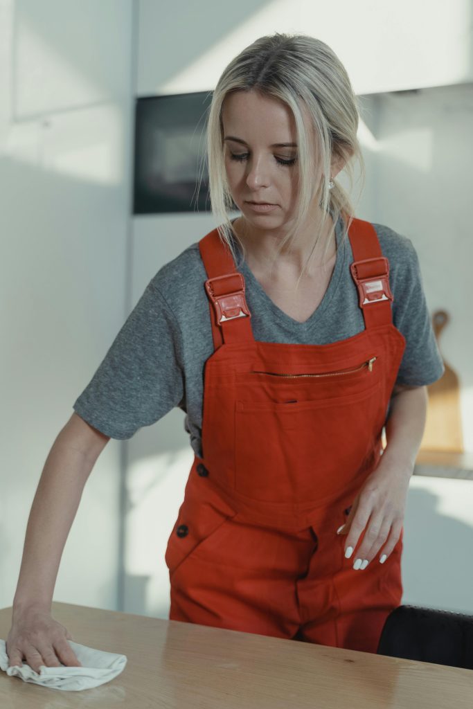 Blonde woman in red overalls sanitizing a wooden table indoors, showcasing home cleaning service.