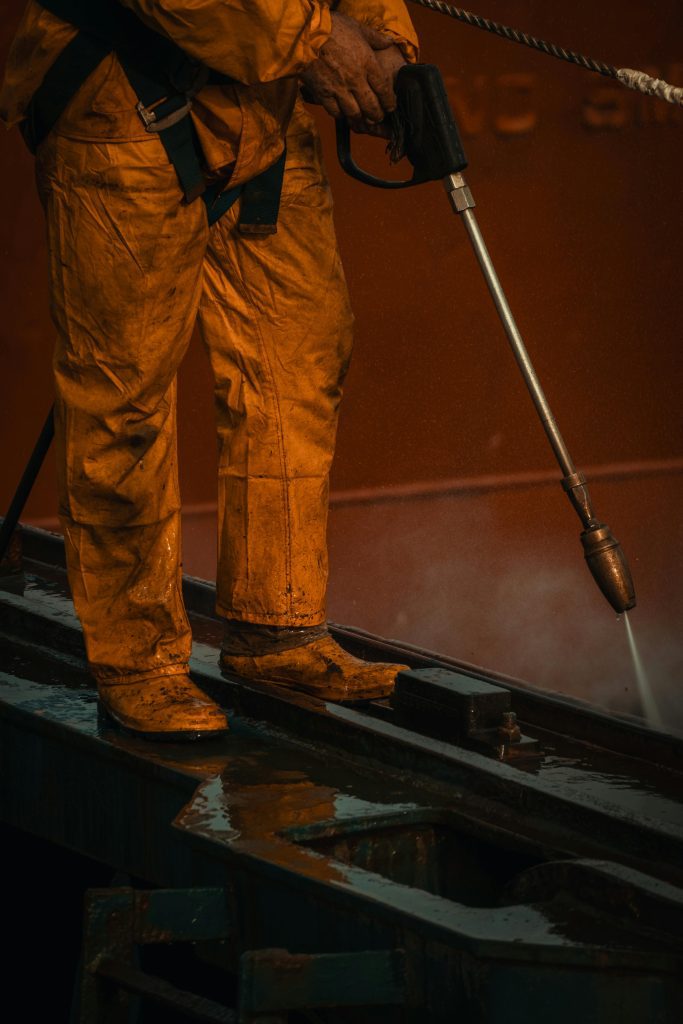 A worker in yellow safety gear uses a pressure washer on industrial equipment.